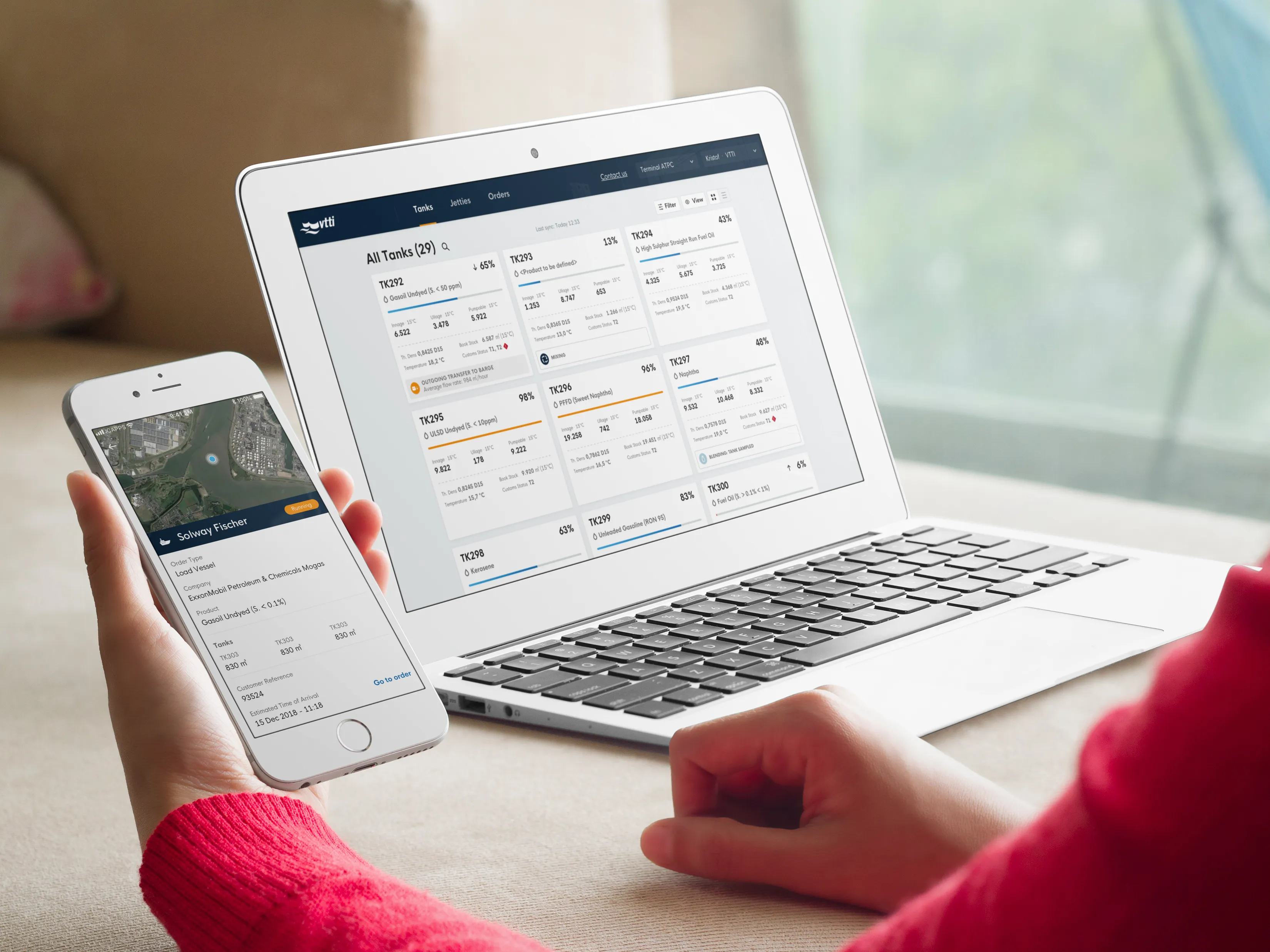 Person holding an iPhone in their hand, where they see the location of a ship. On the laptop next to it is a screen displaying the contents of different tanks on the VTTI terminal